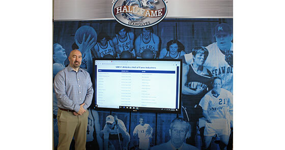 Matt Richards, Associate Dean of Students and Director of Athletics, in front of the interactive Athletics Hall of Fame inside the HUB lobby.